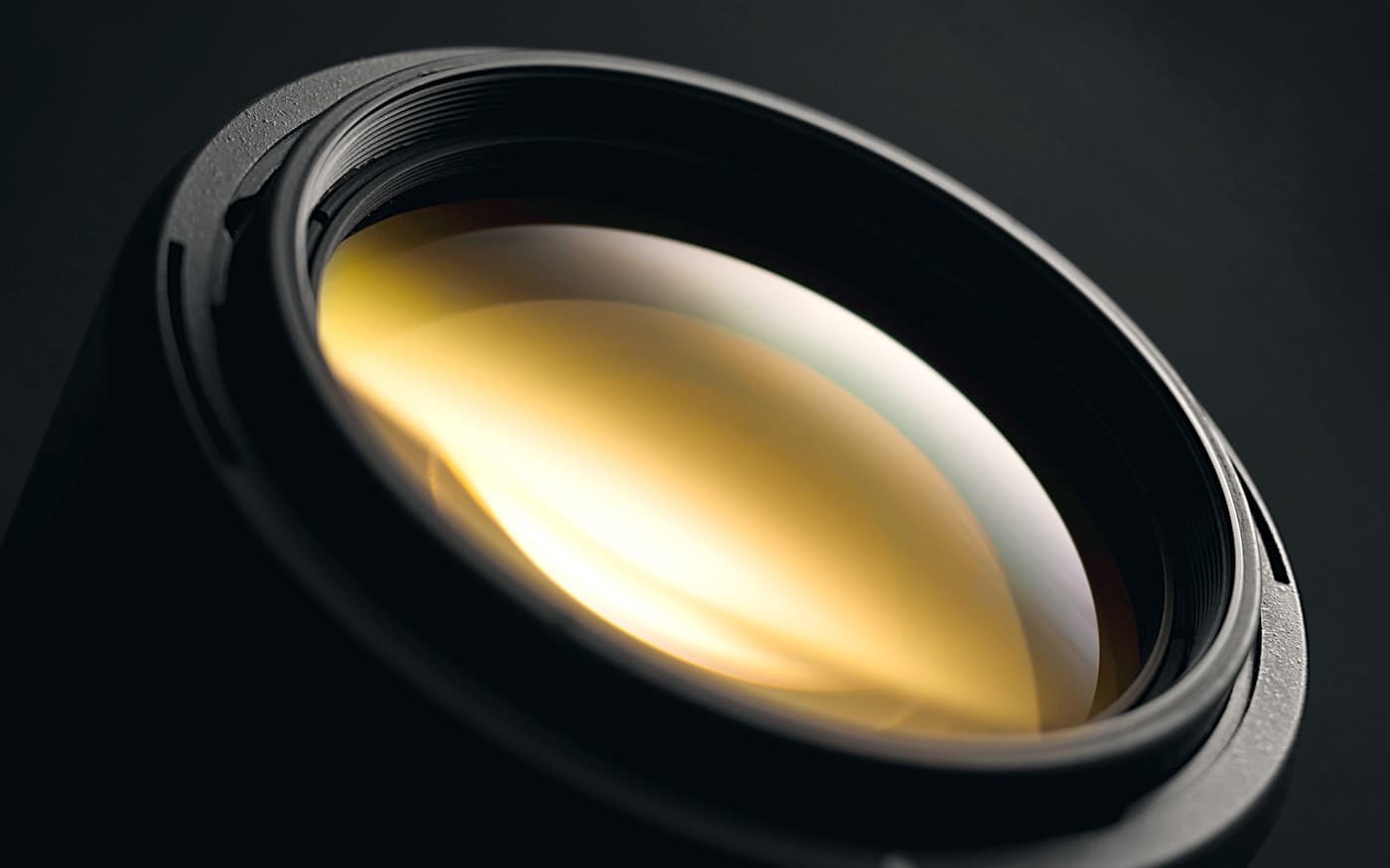 A large lens element on black, a band of warm light across the coating.