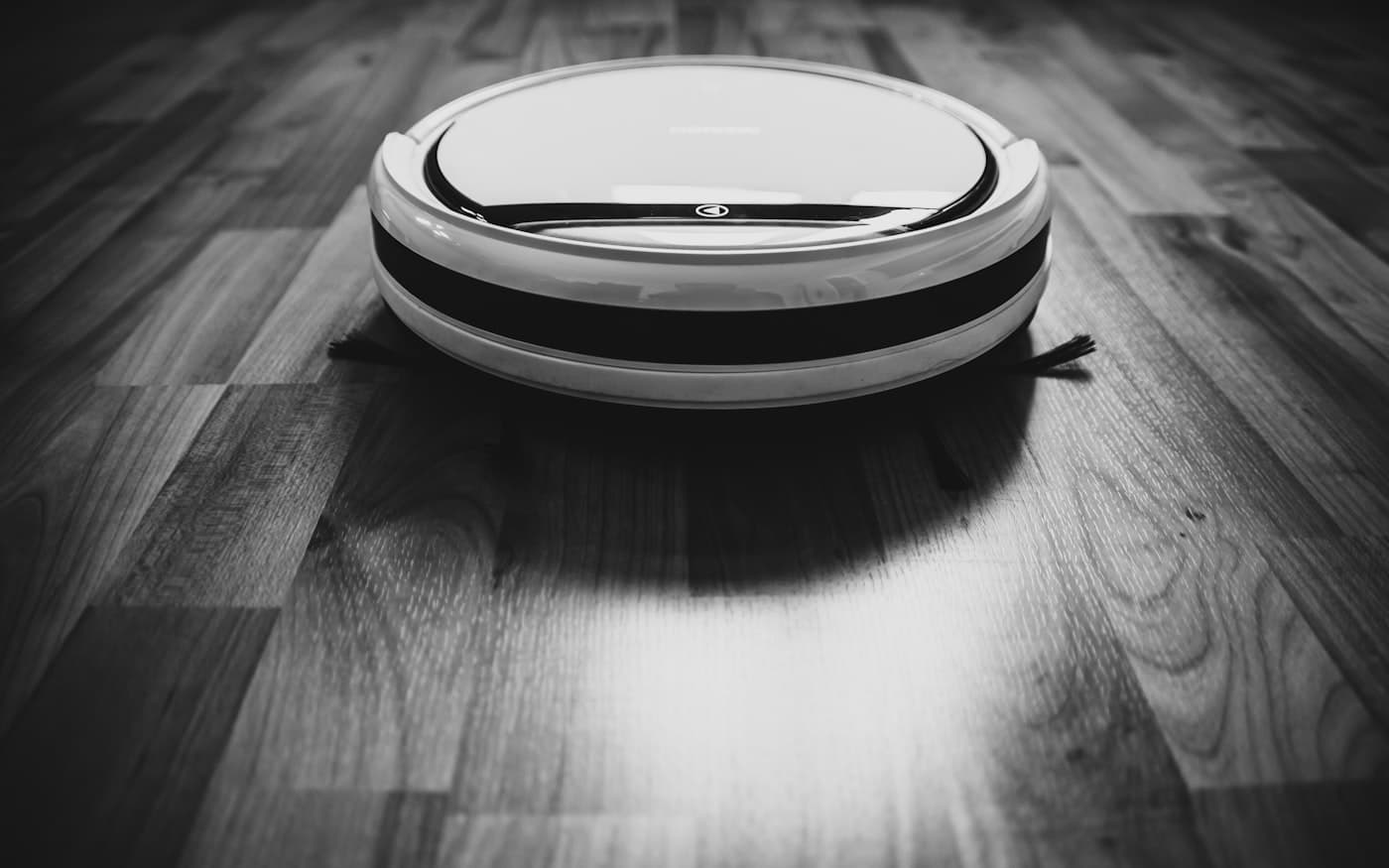 A robot vacuum on a hard floor in black and white, throwing a long shadow.